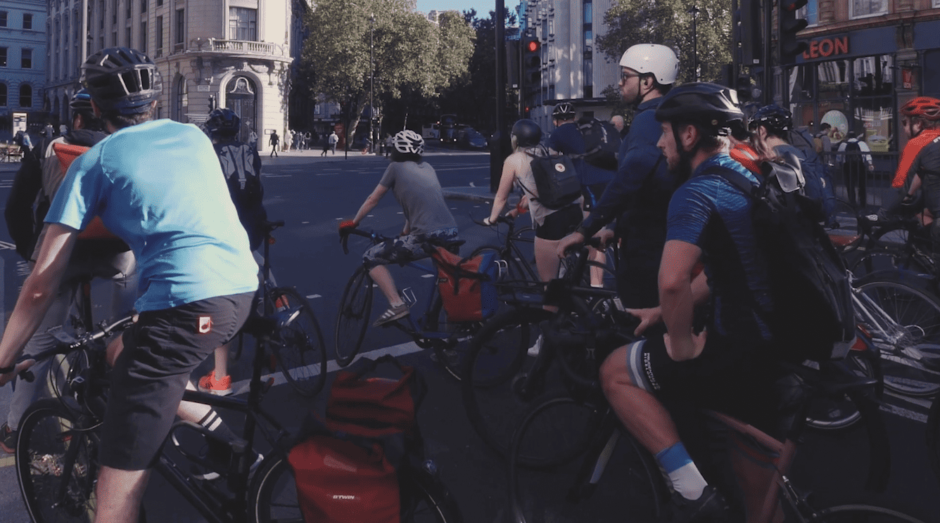 NR: A group of people riding bicycles on a city street while being observed by Norman Rourke Pryme.